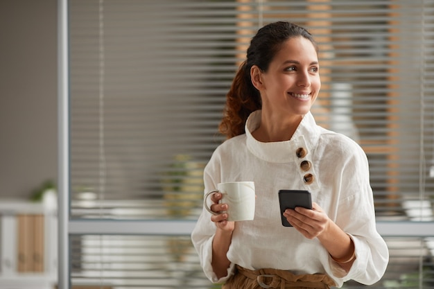 Taille-up portret van lachende elegante vrouw met koffiemok en smartphone terwijl u geniet van pauze van het werk, kopieer ruimte