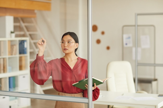 Taille-up portret van moderne Aziatische zakenvrouw schrijven op glazen wand tijdens de planning van project in kantoor, kopieer ruimte