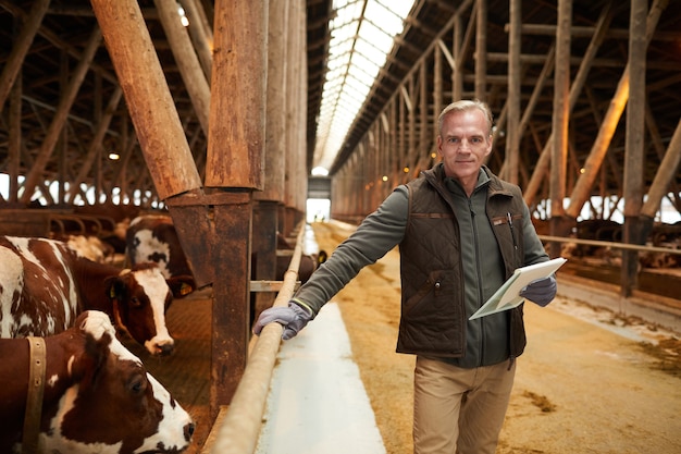 Taille-up portret van moderne volwassen man klembord houden en glimlachen naar de camera tijdens het inspecteren van vee op schuur in melkveebedrijf, kopie ruimte