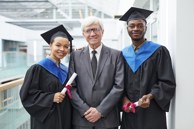 Taille-up portret van twee afgestudeerden met diploma's terwijl ze poseren met een volwassen professor en glimlachen naar de camera