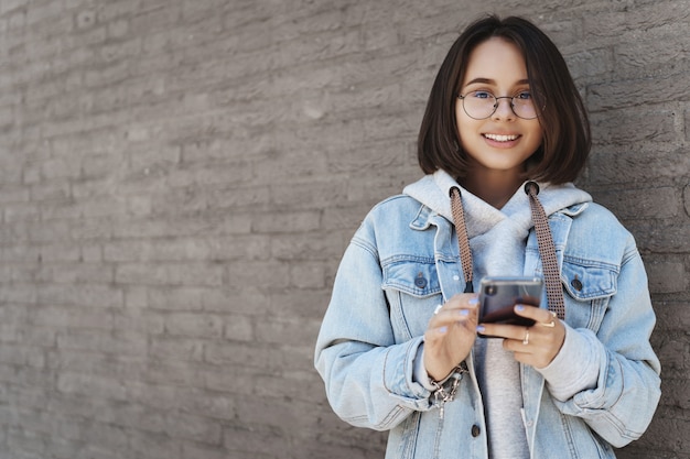 Taille-up portret van vrolijke jonge vrouwelijke student, meisje met behulp van mobiele telefoon op straat, messaging, buiten wachten op vriend, met behulp van kaart-app of sms'en iemand, glimlachend camera in de buurt van bakstenen muur.