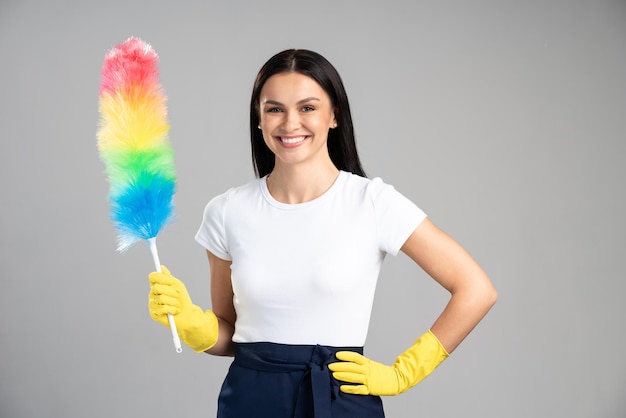 Taille-up portret weergave van de gelukkige dienst schoonmaakster in gele handschoenen met kleur borstel voor stof geïsoleerd op een grijze achtergrond. Huishoudelijk werk en schoonmaak dienstverleningsconcept