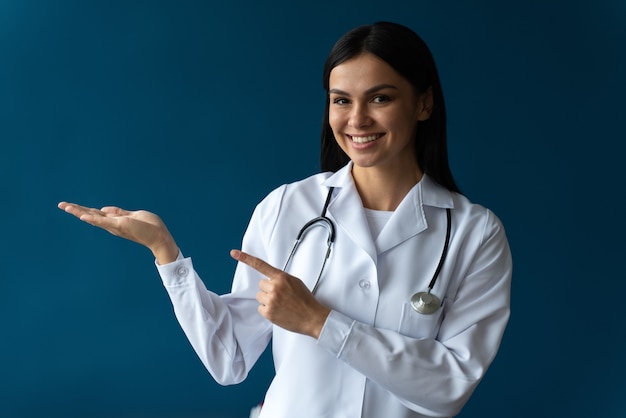 Taille-up portretweergave van de lachende gelukkige dokter wijzend met haar handpalm naar de vrije ruimte op blauwe achtergrond. Arts en geneeskundeconcept