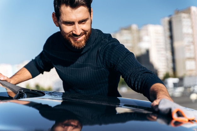 Taille-up portretweergave van een tevreden geconcentreerde man die de achterkant van zijn nieuwe auto schoonmaakt terwijl hij tijd doorbrengt op straat tijdens de zonnige zomerdag Stockfoto