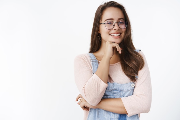 Taille-up shot van charmante, geamuseerde en schattige jonge vrouw met bruin haar in een bril en tuinbroek die zachtjes met de kin op de hand leunt