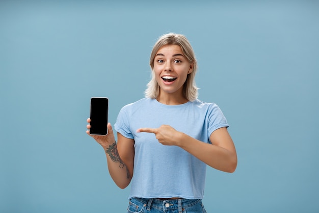 Taille-up shot van opgewonden en onder de indruk knappe vrouwelijke student in casual t-shirt glimlachend vreugdevol wijzend naar smartphonescherm met geweldige plek via internet aan vriend over blauwe muur