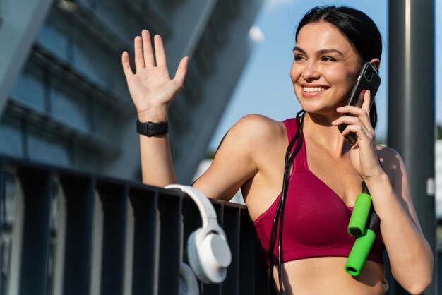 Tailleportret van vrolijke sportvrouw met springtouw en smartphone die naar iemand zwaait