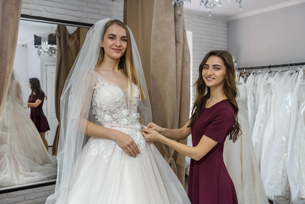 Tailor in wedding salon helping bride to try on  dress