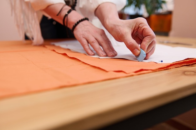 tailor at work closeup of a dressmaker's hands transferring sewing patterns for clothes on orange colored satin fabric in fashion design atelier