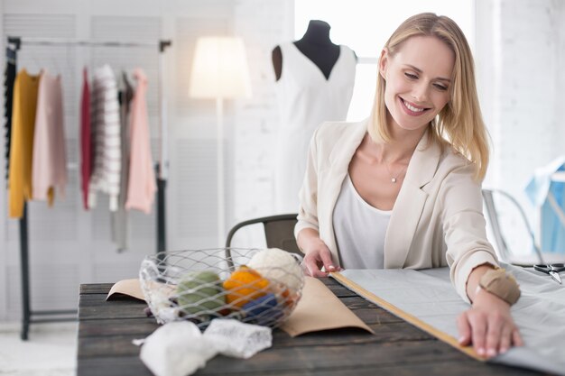 Tailors studio. Jovial female dressmaker working with fabric and applying meter