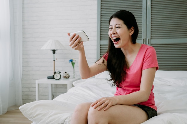 Taiwanese girl sitting at bedside seeing hilarious talk show on\
her smartphone. asian millennial woman is having a good laugh at\
some humorous conversation between two characters in a comedy.