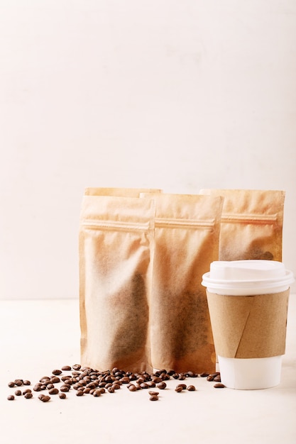 Take away paper cup, coffee beans and coffee packs with blank space for logo over white wall. Copy space