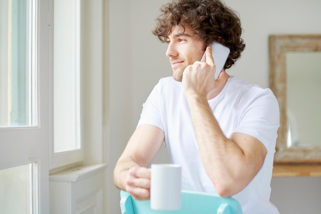 気楽に 自宅の居間に座って携帯電話で話し、コーヒーを飲んでいる幸せな若い男のショット