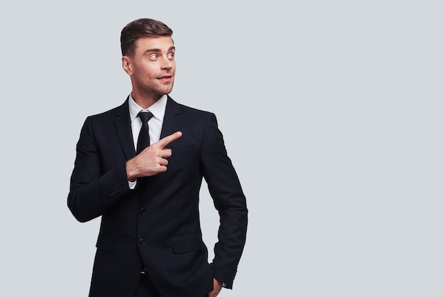 Take a look over there! Good looking young man in full suit pointing copy space and smiling while standing against grey background