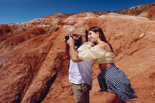 Take photos in open mountain nature. Adult man with a beard in  white shirt and panama hat holds digital camera in his hands and takes pictures of the canyon. The girl leans on him and looks smiling.