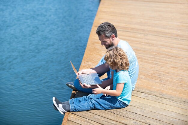 夏休みにeラーニングを受講してください。父と息子は海の桟橋に座ってラップトップを使用しています。ホームスクーリングとオンラインスクーリング。リモート学習。遠隔教育。家族をeラーニングに参加させる。