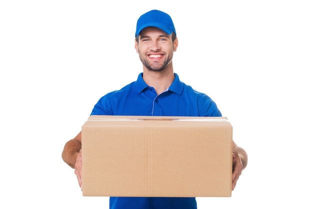 Take your package! Happy young courier stretching out a cardboard boxand smiling while standing against white background