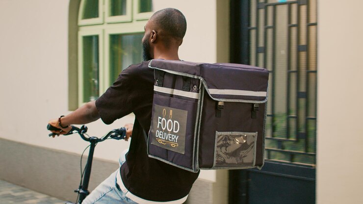 Photo takeaway carrier going to office building entrance, young man working as food delivery courier on bicycle. african american fastfood employee with thermal bag riding bike on street.