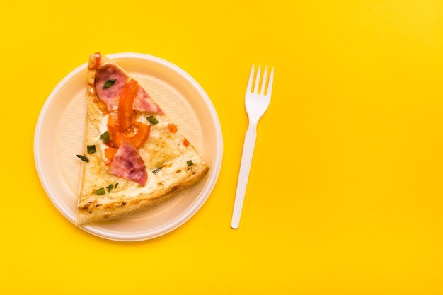Takeaway and delivery pizza slice in disposable plastic plate and fork on yellow background