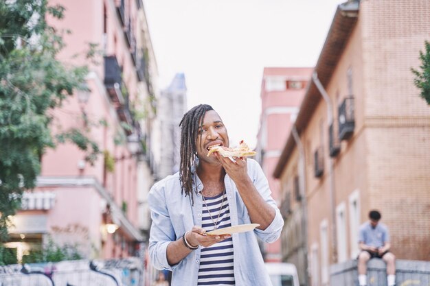 takeaway food man with braids eating a slice of pizza on the street walking