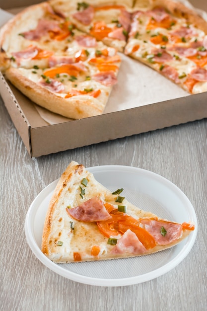 Еда на вынос. Кусок пиццы в одноразовой пластиковой тарелке и коробка пиццы на столе на кухне. Вертикальный вид