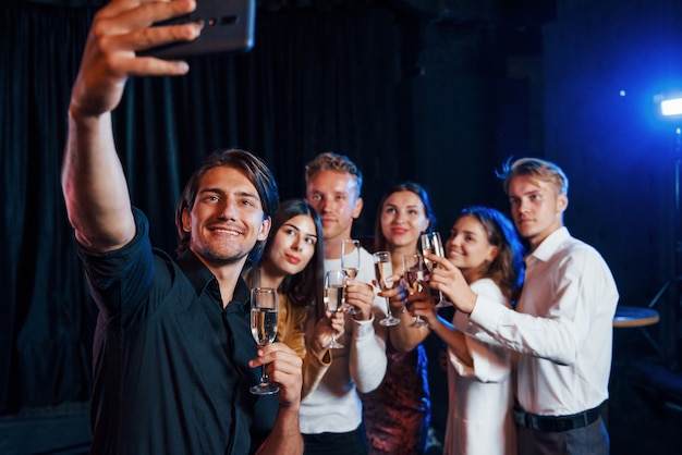 Takes selfie. Group of cheerful friends celebrating new year indoors with drinks in hands.