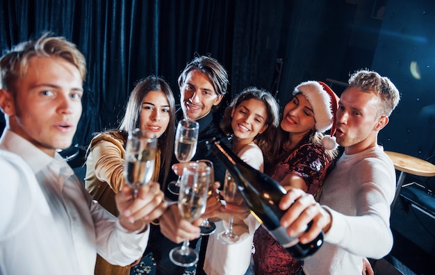 Takes selfie. Group of cheerful friends celebrating new year indoors with drinks in hands.