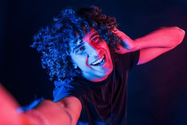 Takes selfie Young beautiful man with curly hair is indoors in the studio with neon lighting