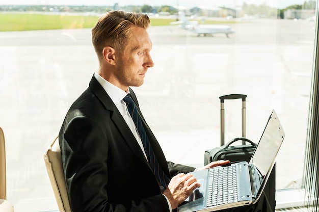 Taking advantages of free Wi-Fi. Side view of confident businessman in formalwear working on laptop while waiting for a flight in airport