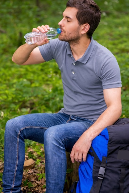 Taking a break after long trip. Tired young man drinking water while sitting in a forest with backpack laying near him with