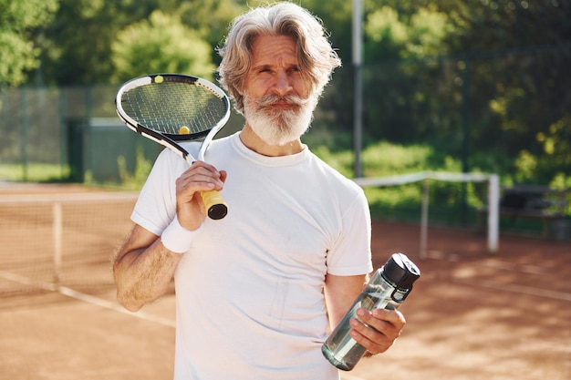 Taking a break and drinking water Senior modern stylish man with racket outdoors on tennis court at daytime