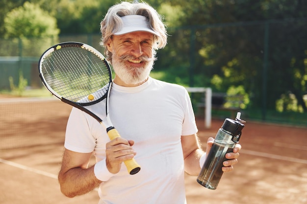 Prendersi una pausa e bere acqua uomo anziano moderno ed elegante con racchetta all'aperto sul campo da tennis durante il giorno
