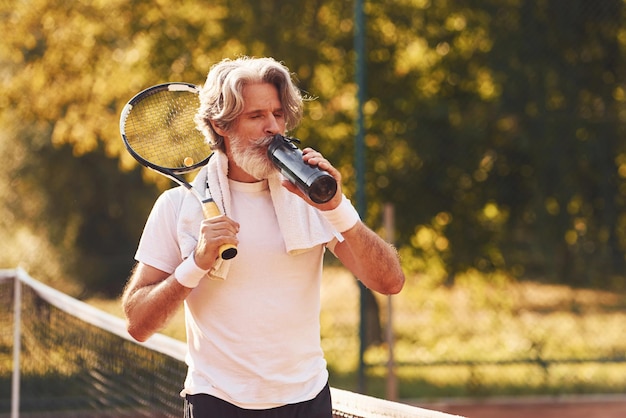 休憩して水を飲むテニスコートで白いシャツと黒いスポーツショーツのシニアスタイリッシュな男