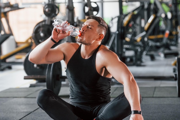 Taking a break and drinking water Young sportive strong man in black wear have workout day in gym