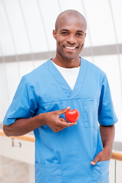 Taking good care of your heart. Confident young African doctor in blue uniform holding heart prop in his hand and smiling