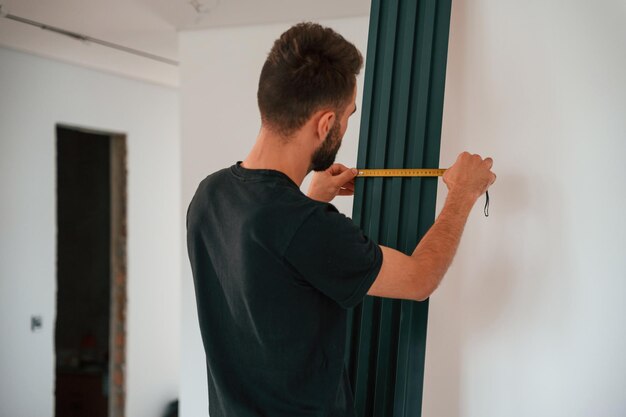 Taking measurements Plumber worker installing heating radiator in empty room of a newly built house