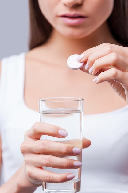 Taking medicines. Close-up of young woman holding glass of water and a pill while standing against grey background