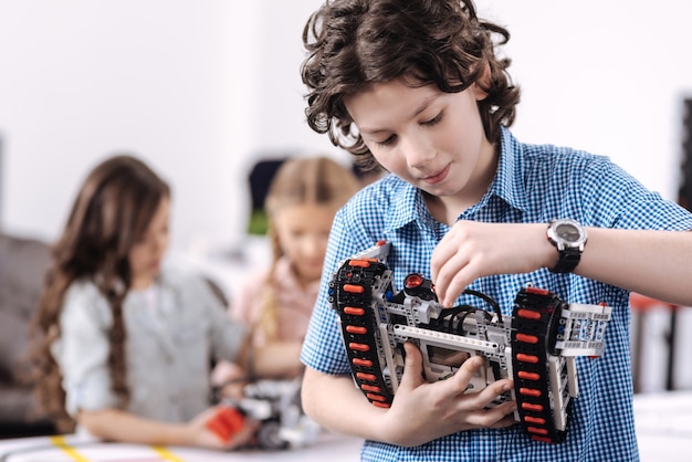 Taking part in science progress. Delighted cheerful concentrated boy standing at school and holding robot while repairing it and his classmates working on the project