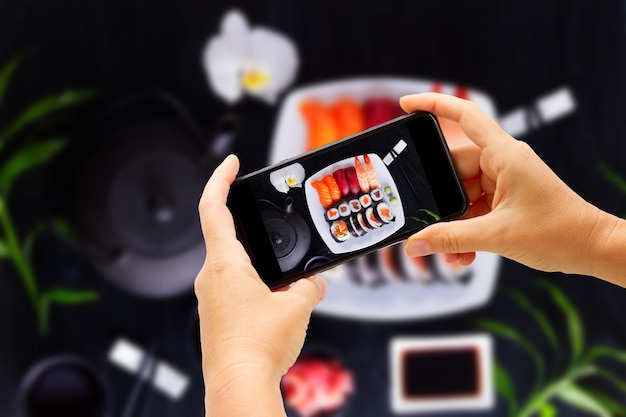 Taking photo of Japanese sushi with teapot and tea cups on black wooden background, top view