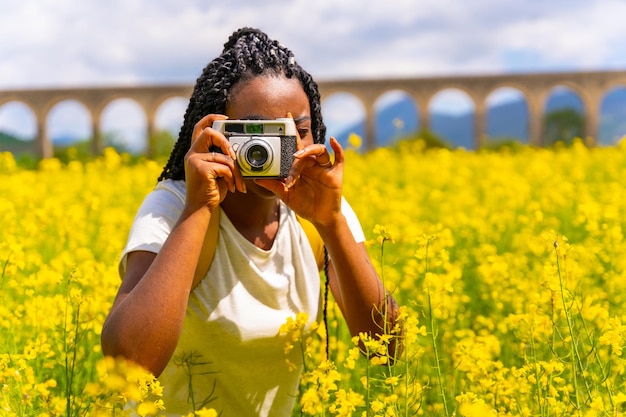 ヴィンテージカメラで写真を撮る黄色い花のフィールドで旅行者を三つ編みの黒い民族の女の子