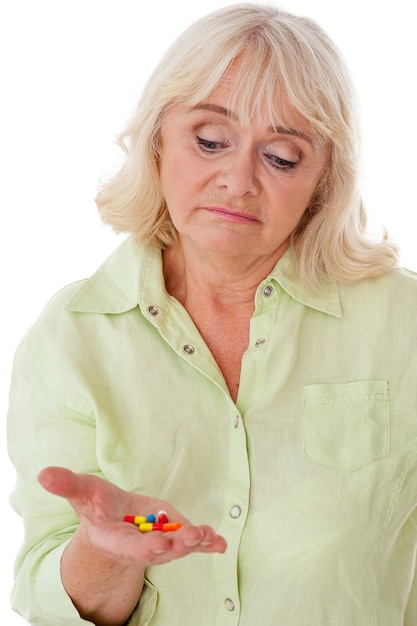 Taking pills. Depressed senior woman holding pills in her hand and looking at them while isolated on white background