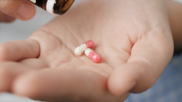 Taking pills. Two small transparent cylindrical capsule tablets with microgranules inside fall into palm of hand from pill bottle. Close-up, front view, center composition