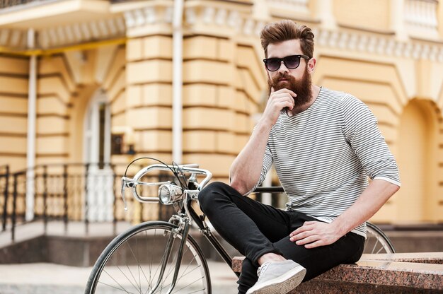 Taking quick break. thoughtful young bearded man holding hand\
on chin and looking at camera while sitting near his bicycle\
outdoors