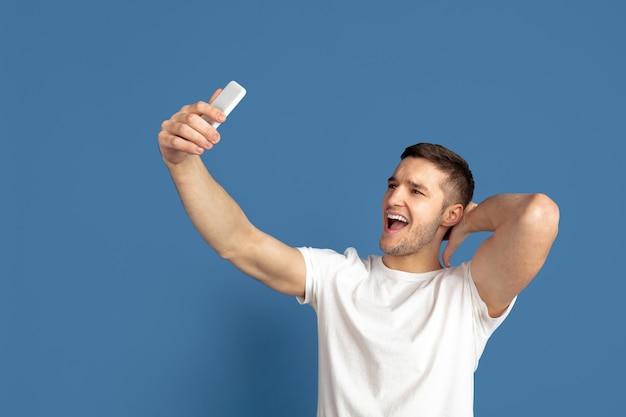 Prendendo selfie. il ritratto del giovane caucasico isolato sulla parete blu dello studio.