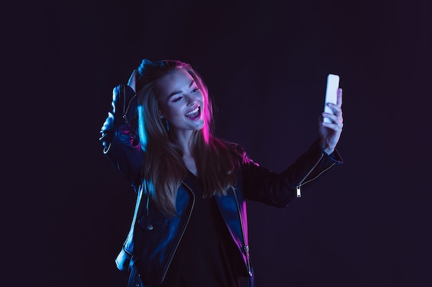 Taking selfie, smiling. Portrait of young beautiful woman in neon light on dark studio backgound. The human emotions, black friday, cyber monday, purchases, sales, finance concept. Copyspace.