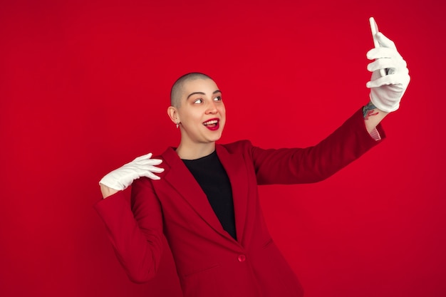 Taking selfie or vlog. Portrait of young caucasian bald woman isolated on red wall.