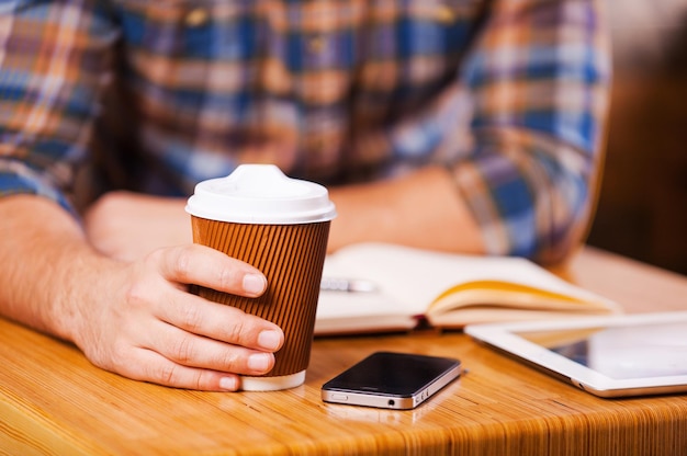 커피 브레이크 시간을 가집니다. 메모장과 디지털 태블릿이 놓여 있는 책상에 앉아 있는 동안 커피 컵을 들고 있는 남자의 클로즈업