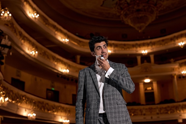 Talented Actor In Suit Wiping Sweat From Face And Forehead With Napkin After Performance, Tired And Worried. Caucasian European Male Standing On Stage. Handsome Brunette Guy In Gray Tuxedo