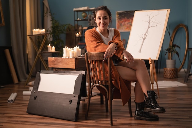 才能あふれるアーティストは、アートスタジオのフォルダー付きの椅子に座って情熱的に微笑んでいます。