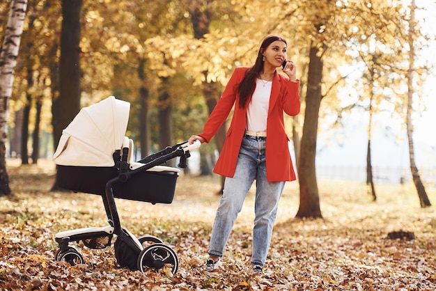 Talking by the phone. Mother in red coat have a walk with her kid in the pram in the park at autumn time.